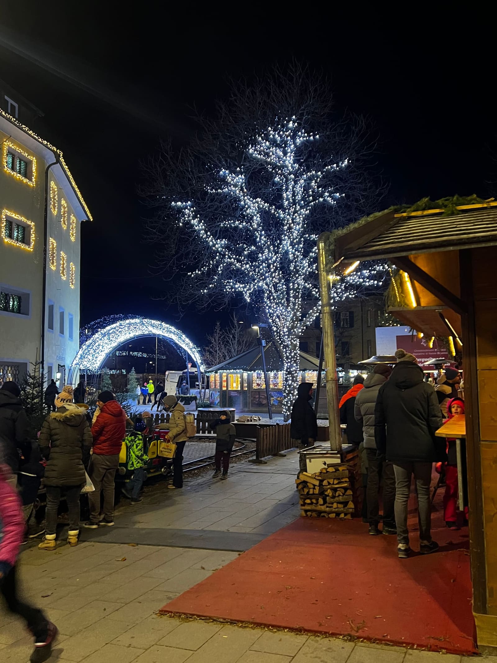 il mercatino di notte, l'arco luminoso all'ingresso e l'albero acceso di bianco