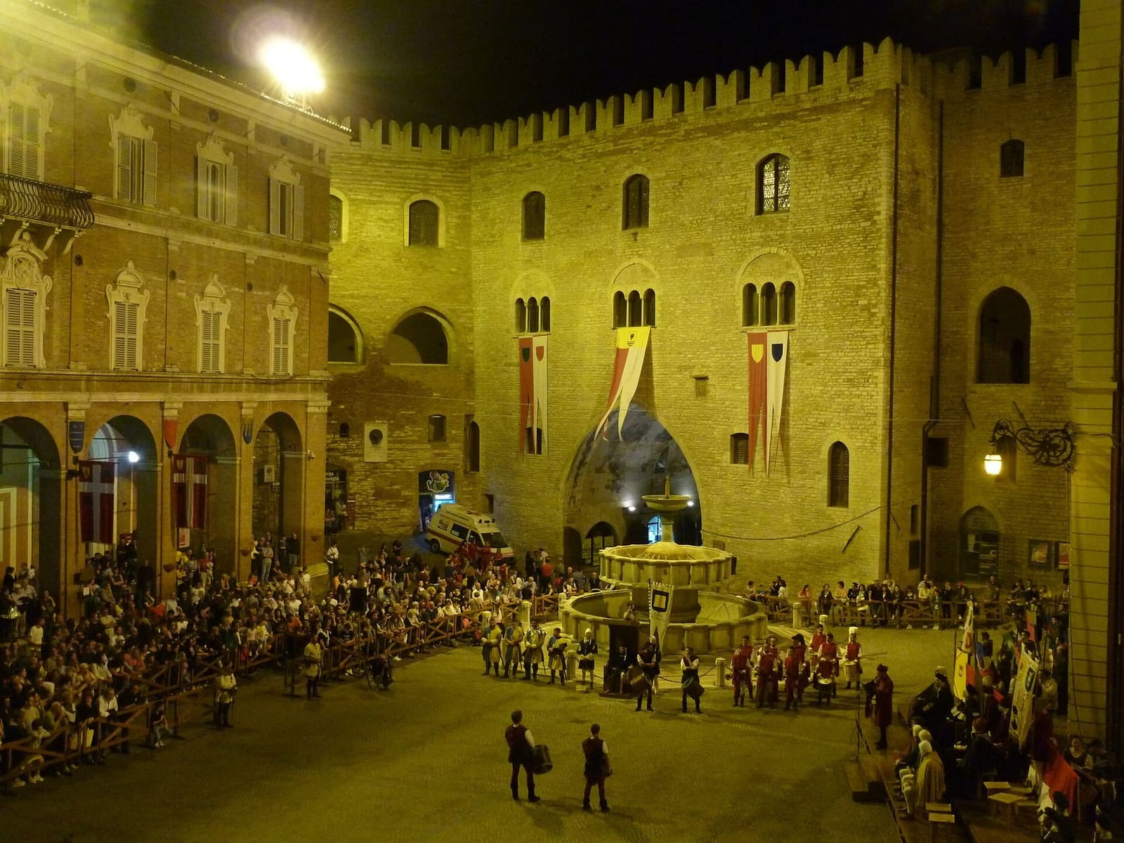 la piazza la sera del Palio di San Giovanni Battista, con i gonfaloni appesi al Podestà e i tamburini in costume intorno alla fontana