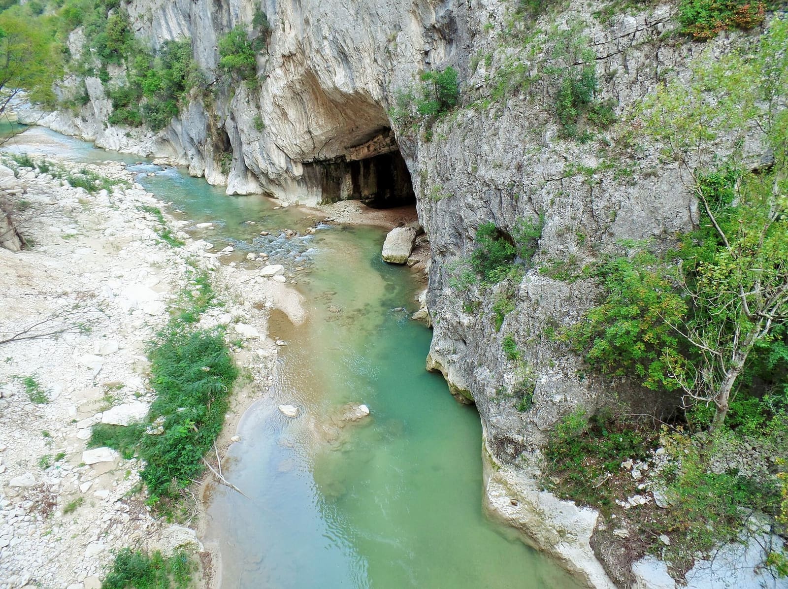 il fiume verde chiaro sotto la parete di calcare, con una grotta che si apre a pelo d'acqua, nel parco della Gola della Rossa e di Frasassi