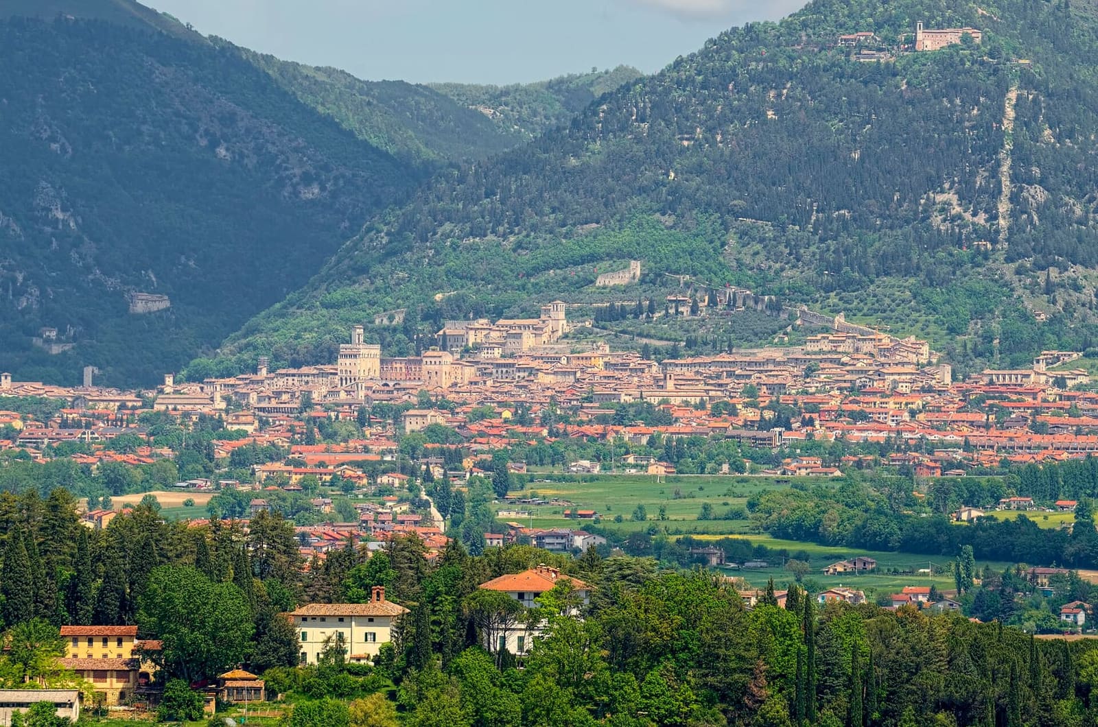 Gubbio da lontano, distesa ai piedi del monte Ingino, con la linea della funivia che sale dritta fino alla basilica