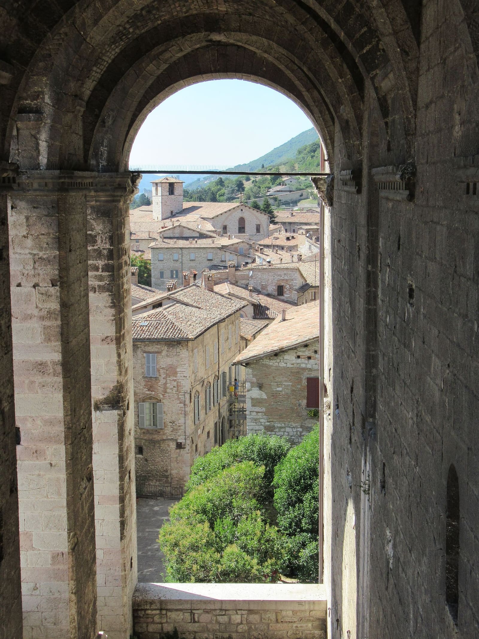 i tetti della città e un campanile inquadrati da un arco del Palazzo dei Consoli, con il fianco del monte sullo sfondo