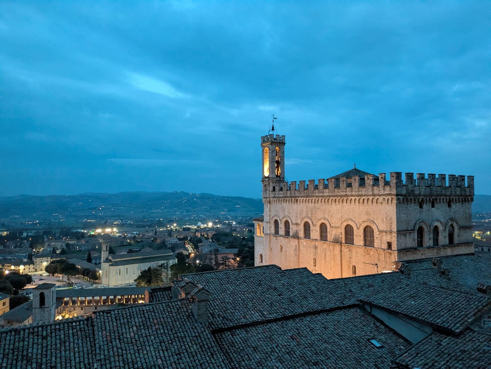 il Palazzo dei Consoli all'imbrunire, acceso sopra i tetti mentre nella pianura si accendono le luci