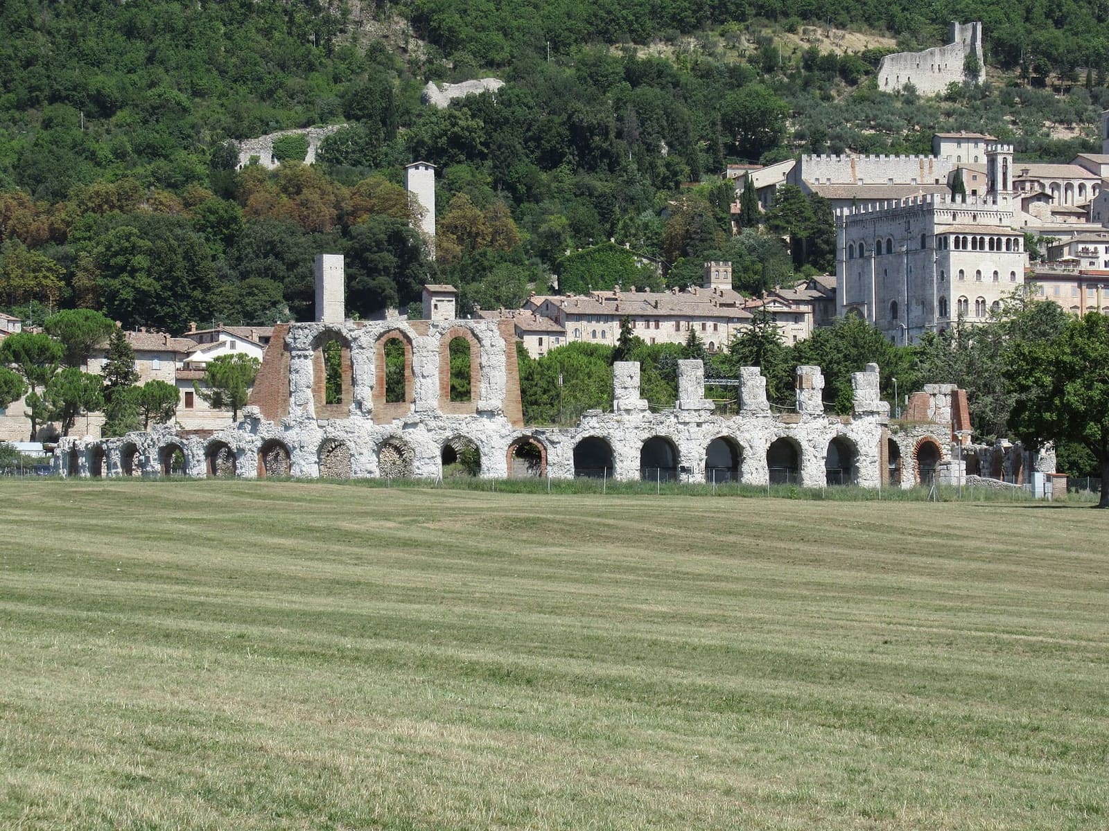 le arcate del teatro romano nel prato della piana, con la città e il Palazzo dei Consoli che salgono sul monte dietro