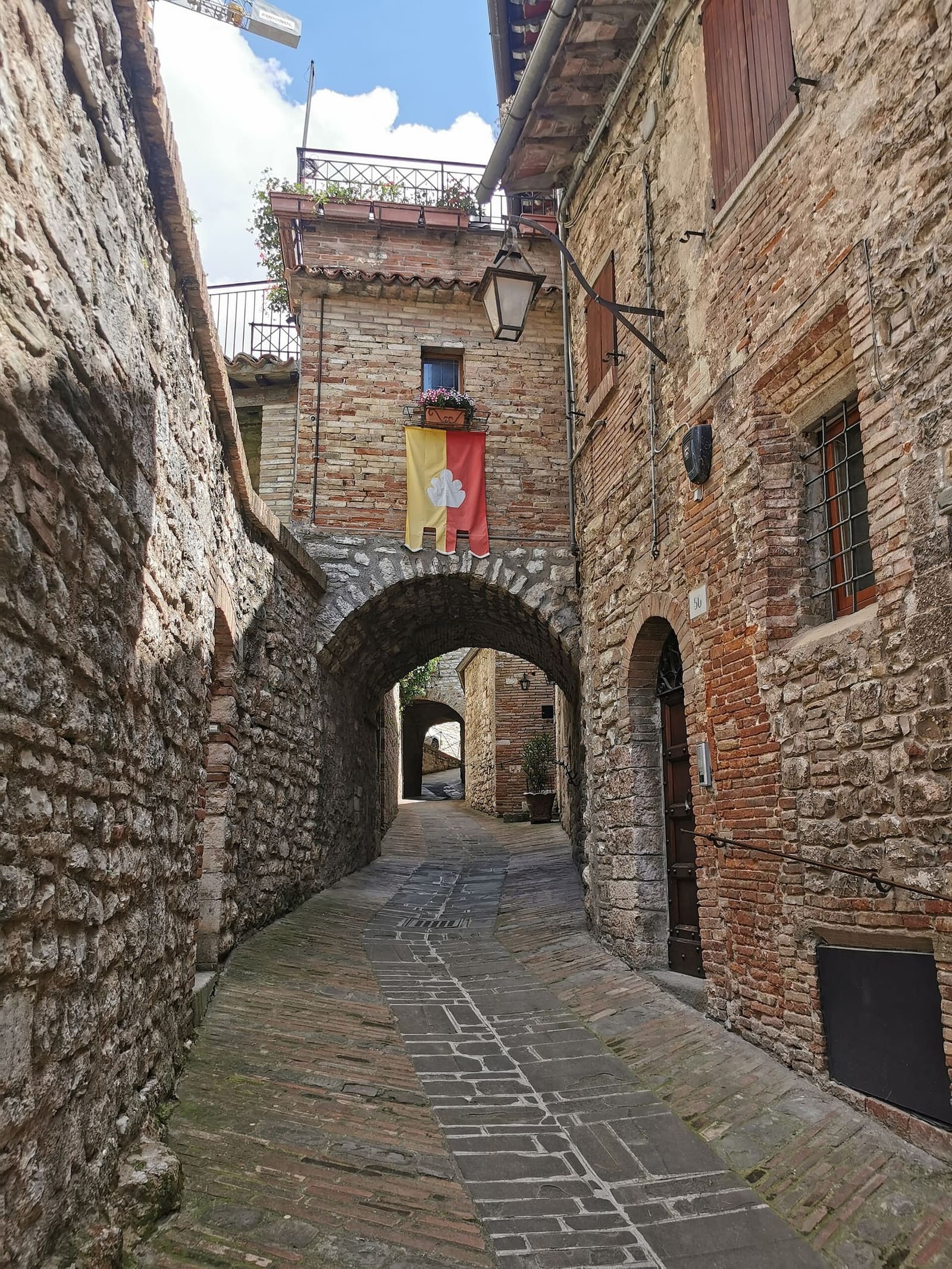un vicolo lastricato che passa sotto un arco di pietra, con il vessillo di un quartiere appeso alla casa in fondo