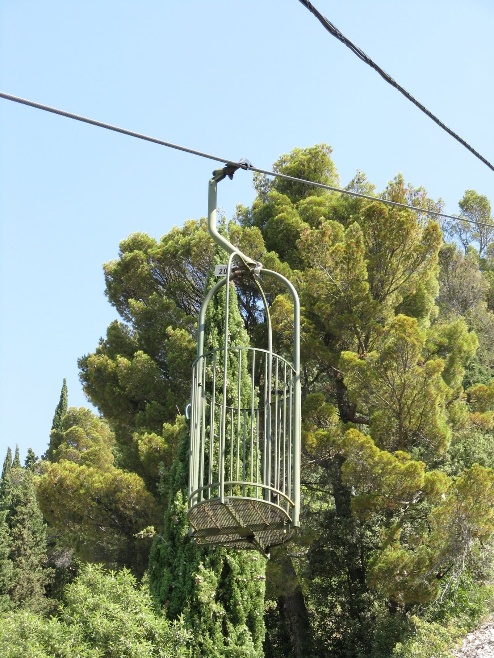una gabbia della funivia Colle Eletto appesa al cavo, aperta e senza porte, fra i pini del monte