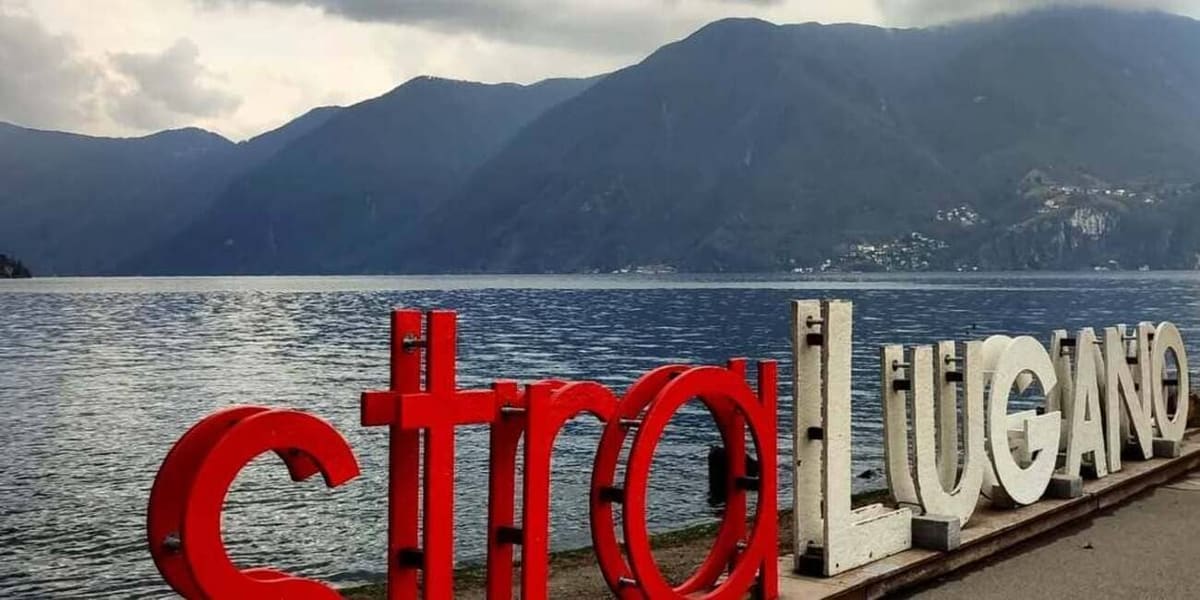 la grande scritta ciao Lugano in lettere rosse e bianche sulla riva, con il lago e le montagne dietro
