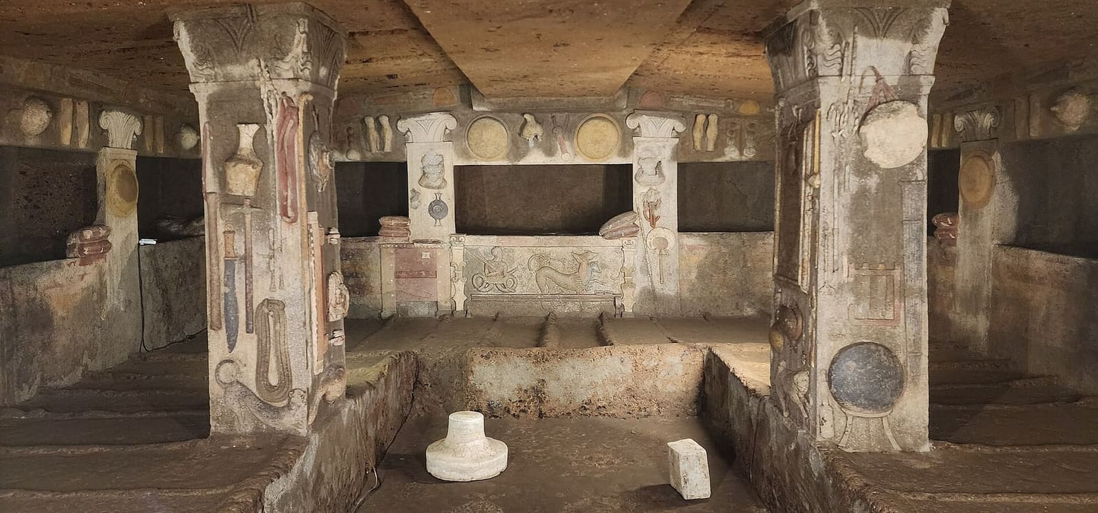 dentro la tomba dei Rilievi: sui pilastri gli attrezzi di casa modellati a stucco e dipinti, una brocca, un coltello, una corda arrotolata, gli scudi rotondi in alto, i cuscini rossi sui letti e in fondo la nicchia del letto grande con Cerbero sotto