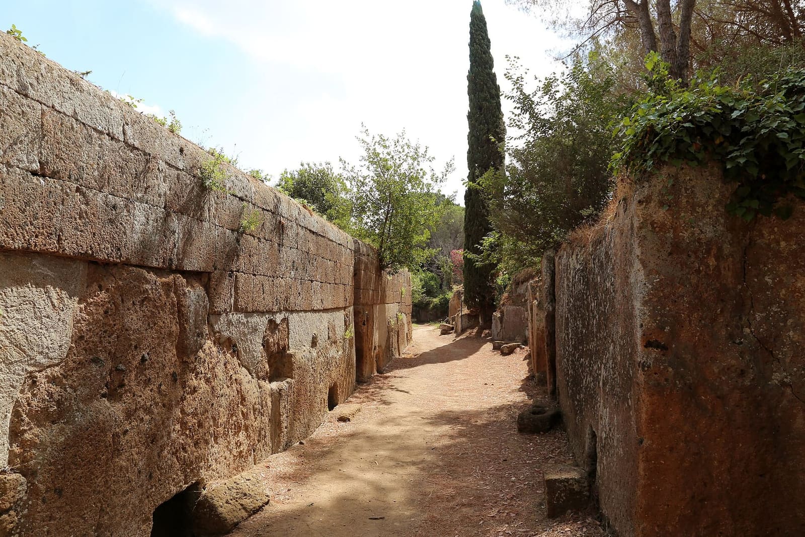 una via stretta fra le pareti di tufo delle tombe a dado, con il sentiero di terra in mezzo e un cipresso in fondo