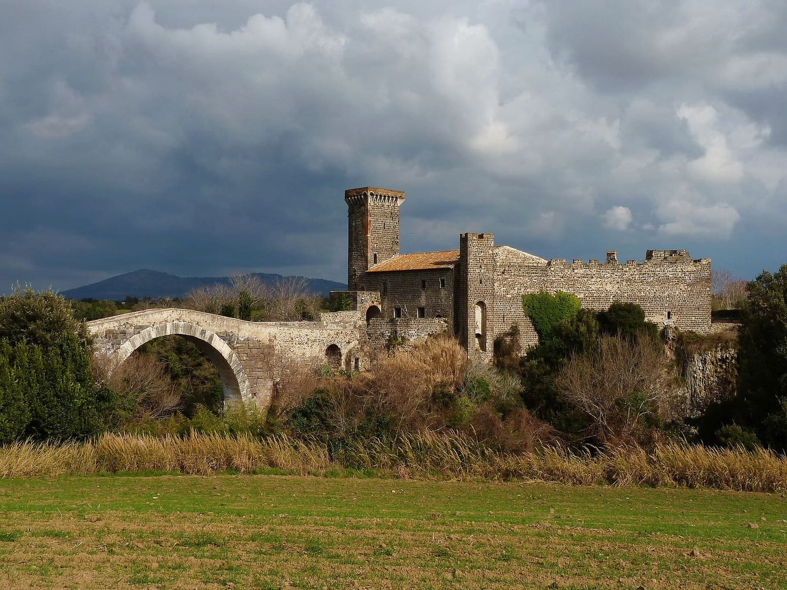 il castello e il ponte dal prato davanti, sotto un cielo di temporale, con i monti scuri sullo sfondo