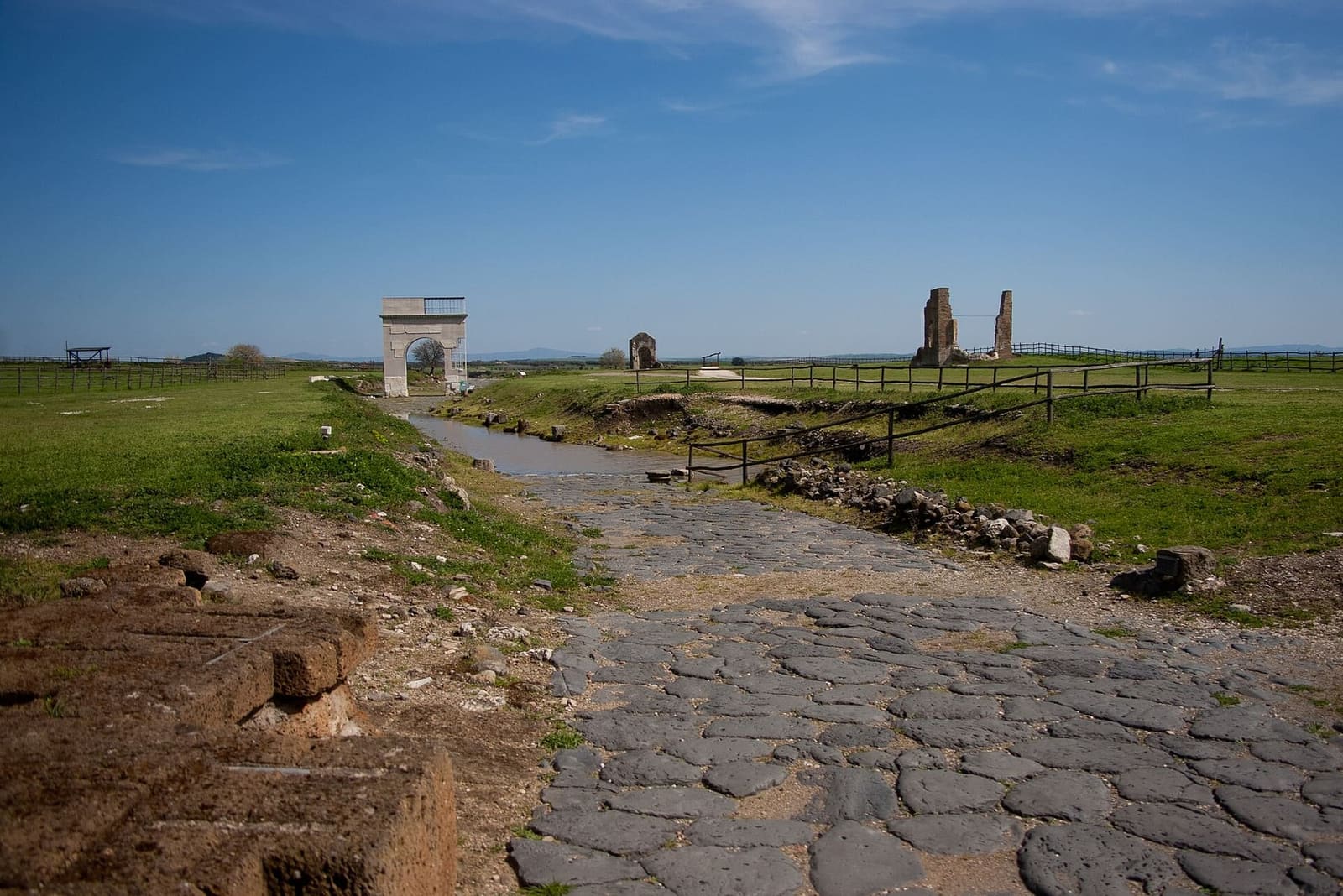 il decumano che attraversa la città, con il basolato di pietra scura in mezzo al prato, l'arco onorario in fondo e a destra i muri di mattoni della basilica