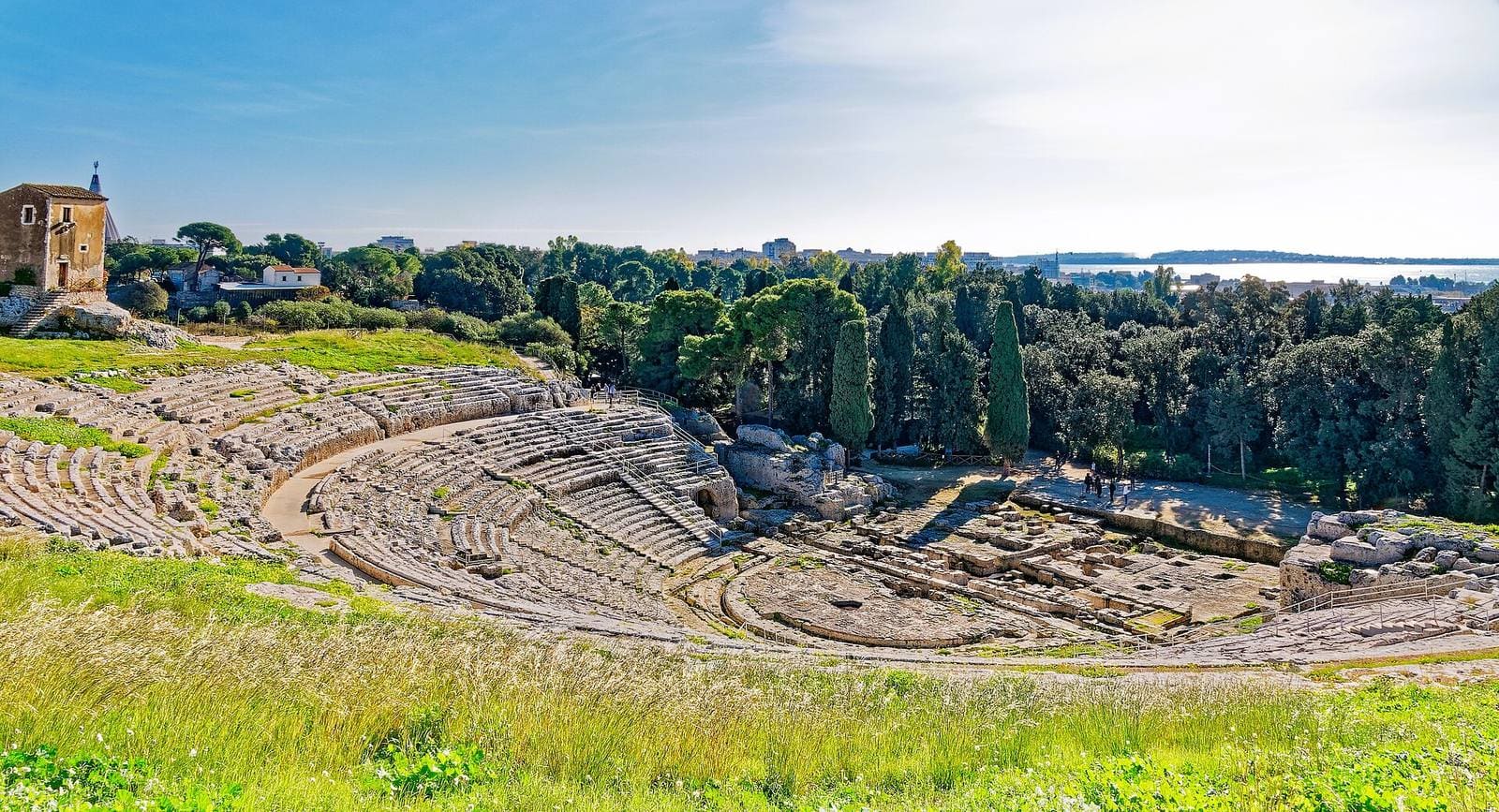la cavea del Teatro greco dall'alto delle gradinate, con la casa dei mugnai a sinistra e il Porto Grande in fondo, oltre i pini