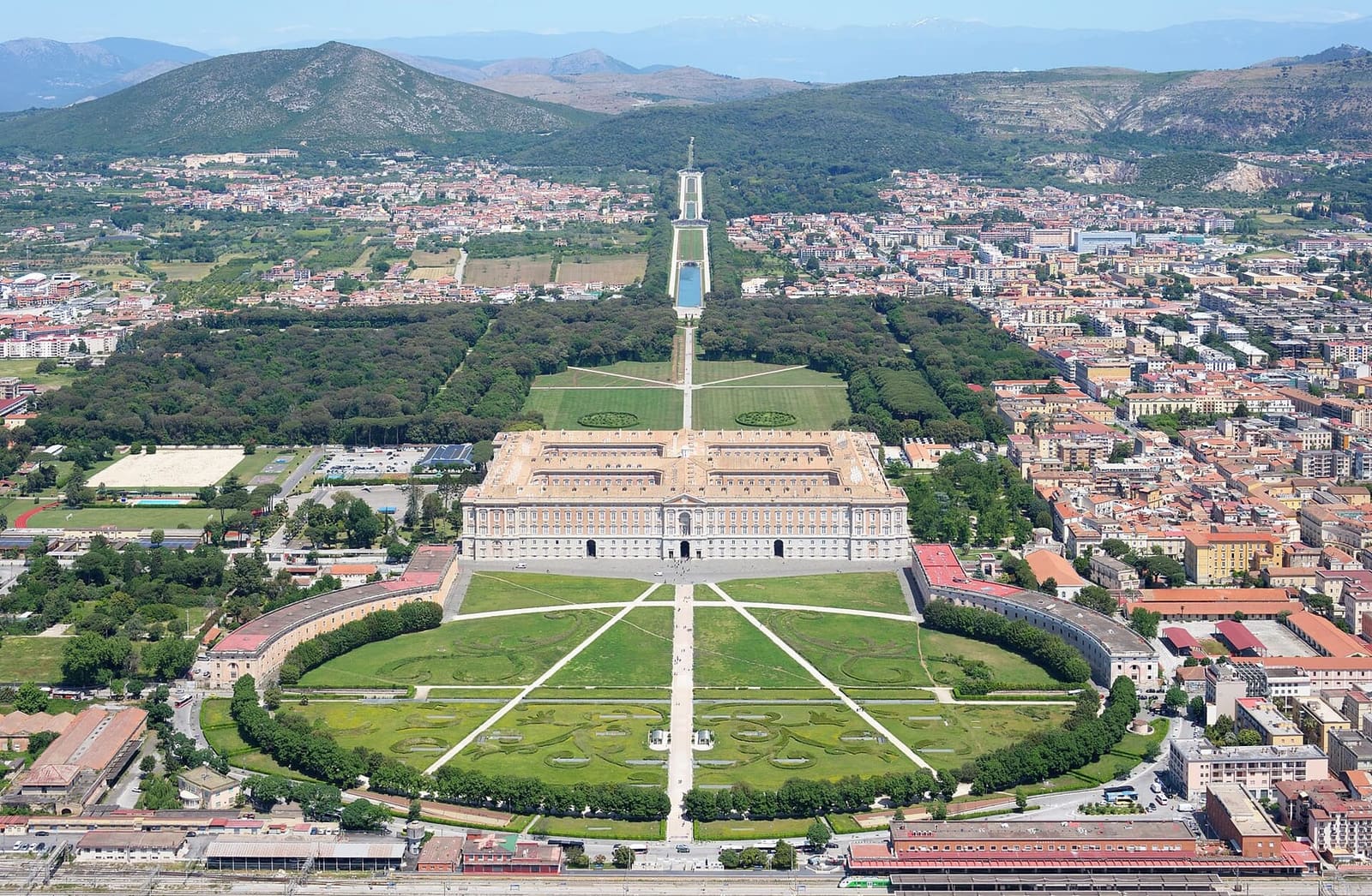 la Reggia vista dall'alto, con la piazza a emiciclo e la stazione davanti e il Parco che sale fino alla cascata