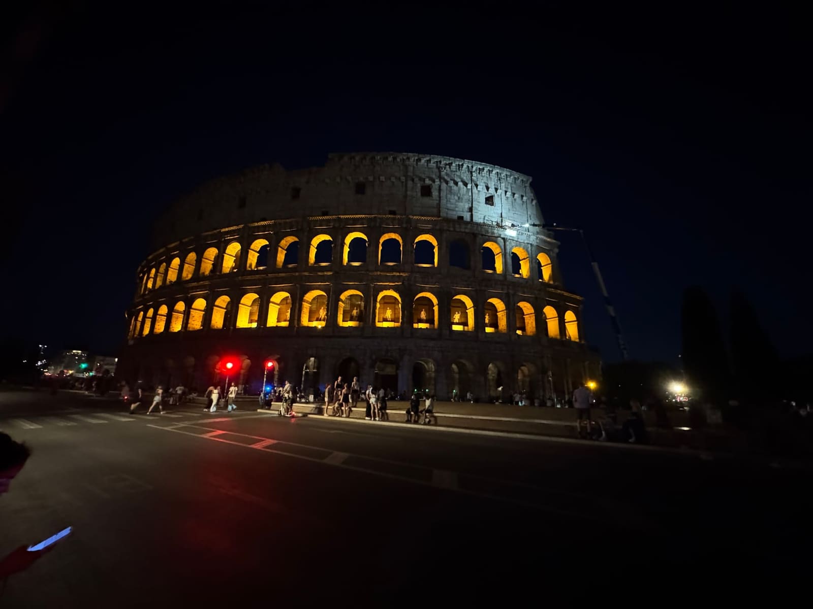 il Colosseo illuminato di sera, visto dalla strada che gli passa davanti, con il semaforo rosso e la gente seduta sul marciapiede