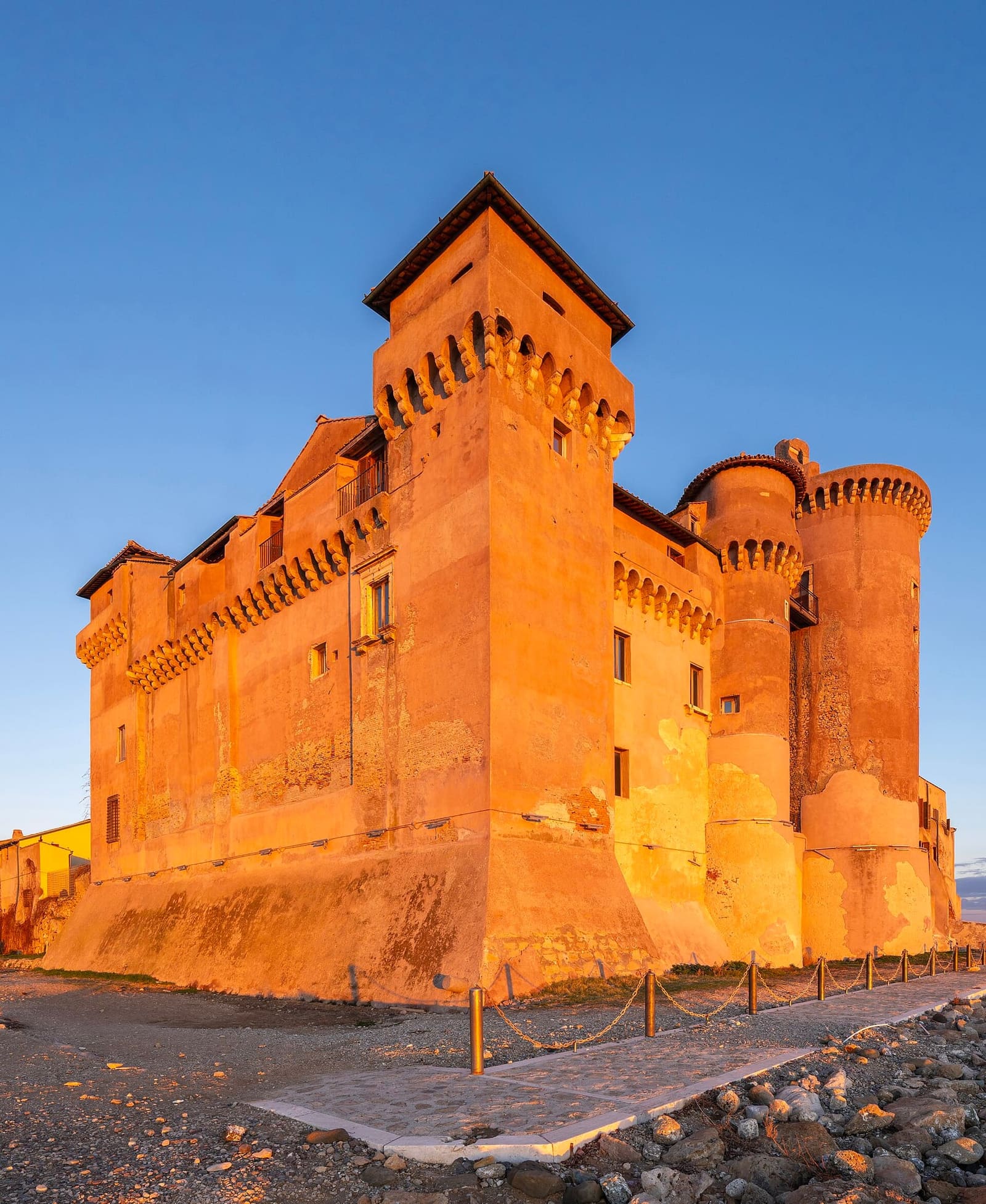 il castello acceso dall'ultimo sole: la torre quadrata con i beccatelli, le due torri rotonde a destra e il camminamento con la catena lungo la riva