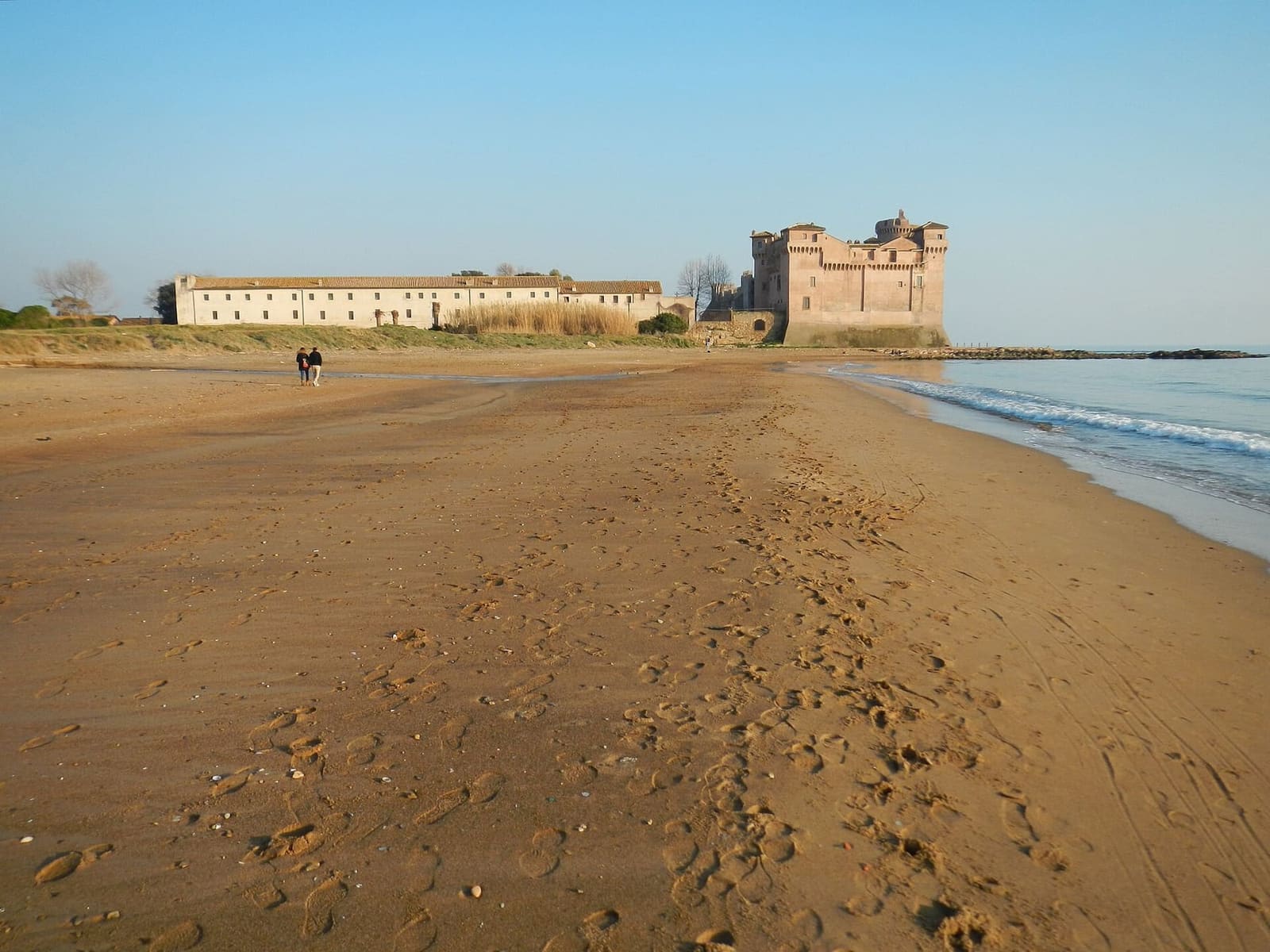 il castello e il lungo edificio del borgo visti dalla spiaggia a ovest, con la sabbia piena di impronte e il mare a destra