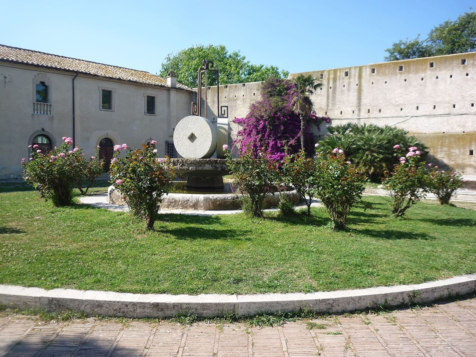 un cortile dentro le mura, con la fontana della macina di pietra in mezzo alle rose e la buganvillea contro il muro di cinta