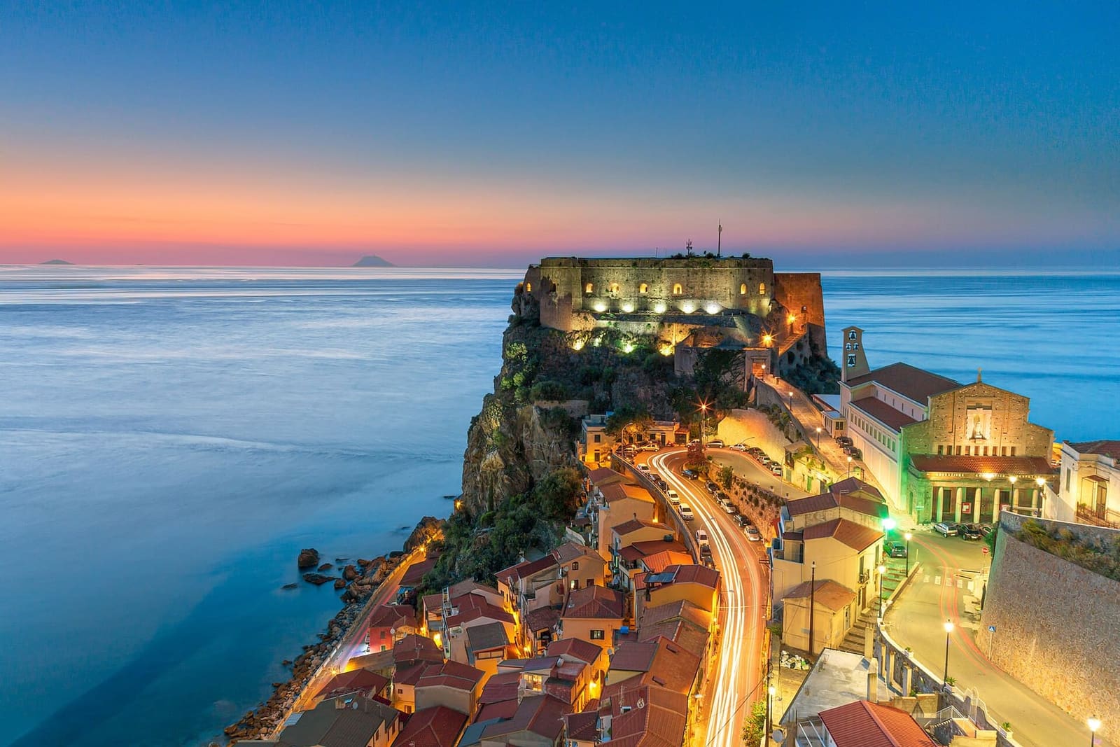 il Castello Ruffo illuminato la sera, con le luci del paese e il mare