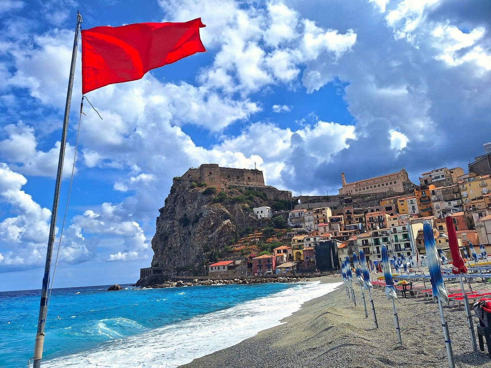 la spiaggia con gli ombrelloni chiusi e la bandiera rossa, e la rupe in fondo