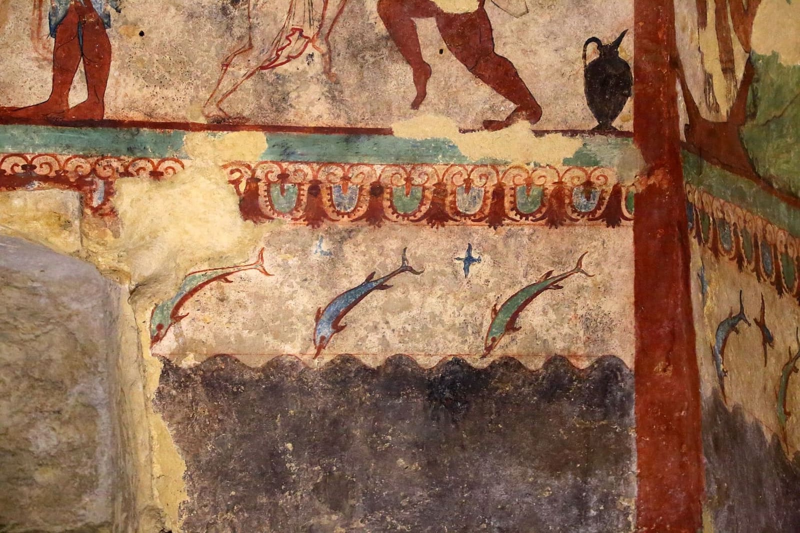 un angolo della Tomba delle Leonesse: i delfini che saltano sopra il mare a onde, la fascia di palmette, una colonna rossa dipinta e una brocca nera appoggiata