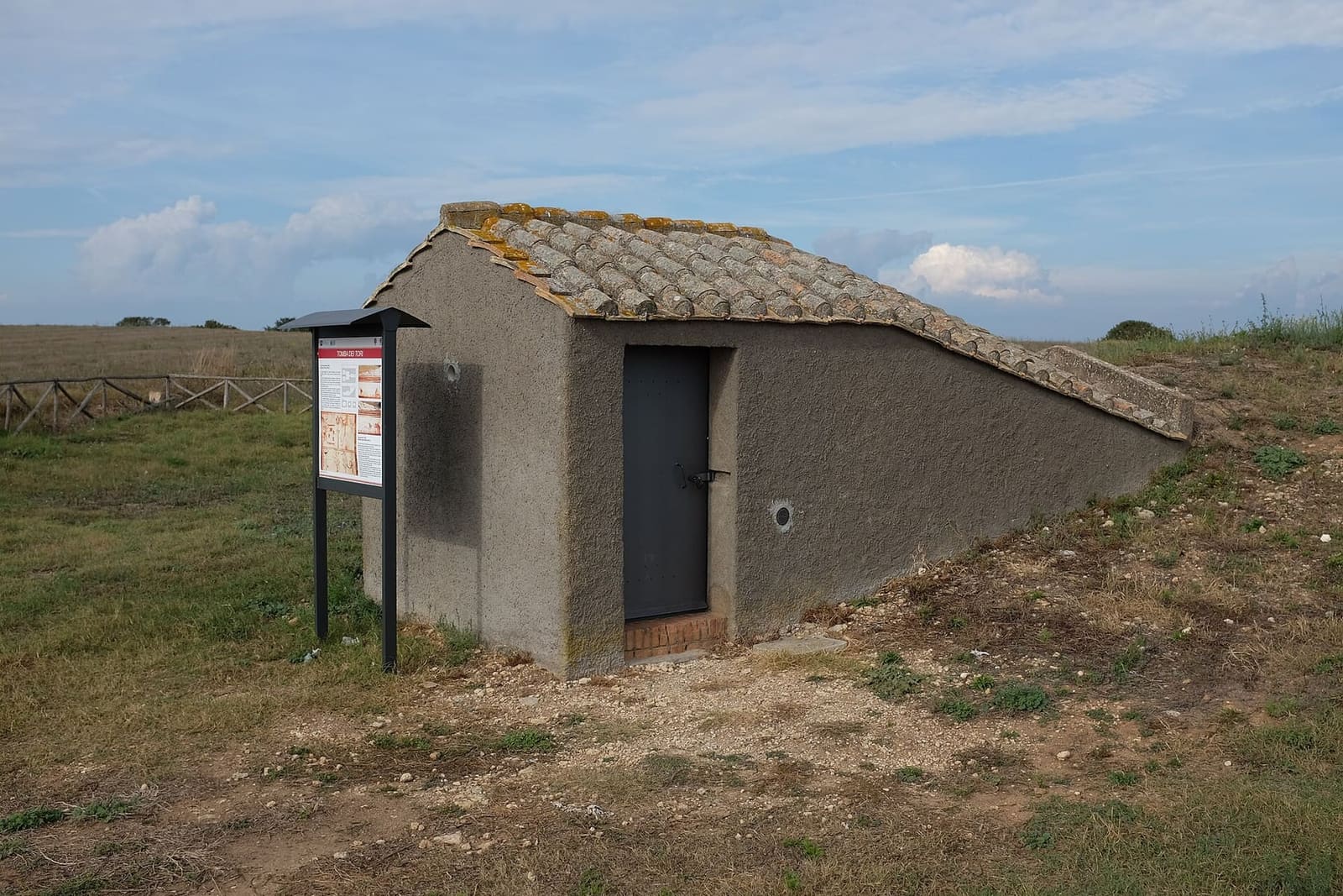il casotto della Tomba dei Tori, con la porta chiusa, il cartello accanto e il tetto di coppi che scende verso il prato: dentro c'è la scaletta