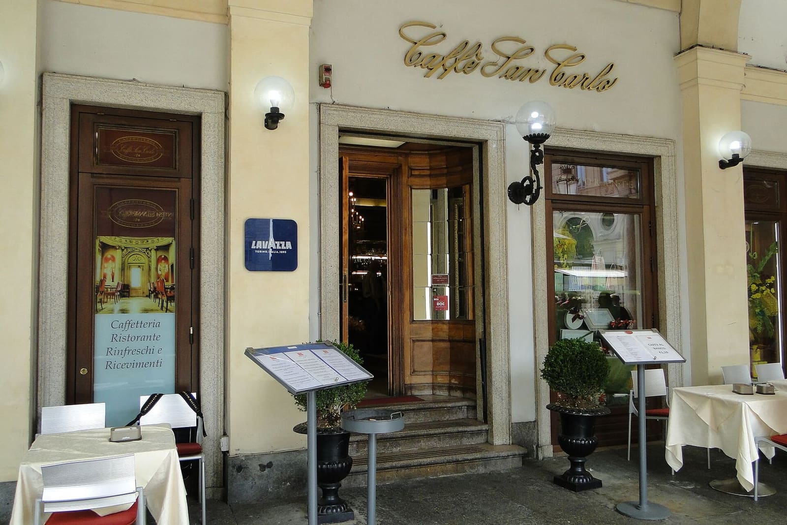 l'ingresso del Caffè San Carlo sotto i portici della piazza, con i tavolini apparecchiati davanti alle porte di legno