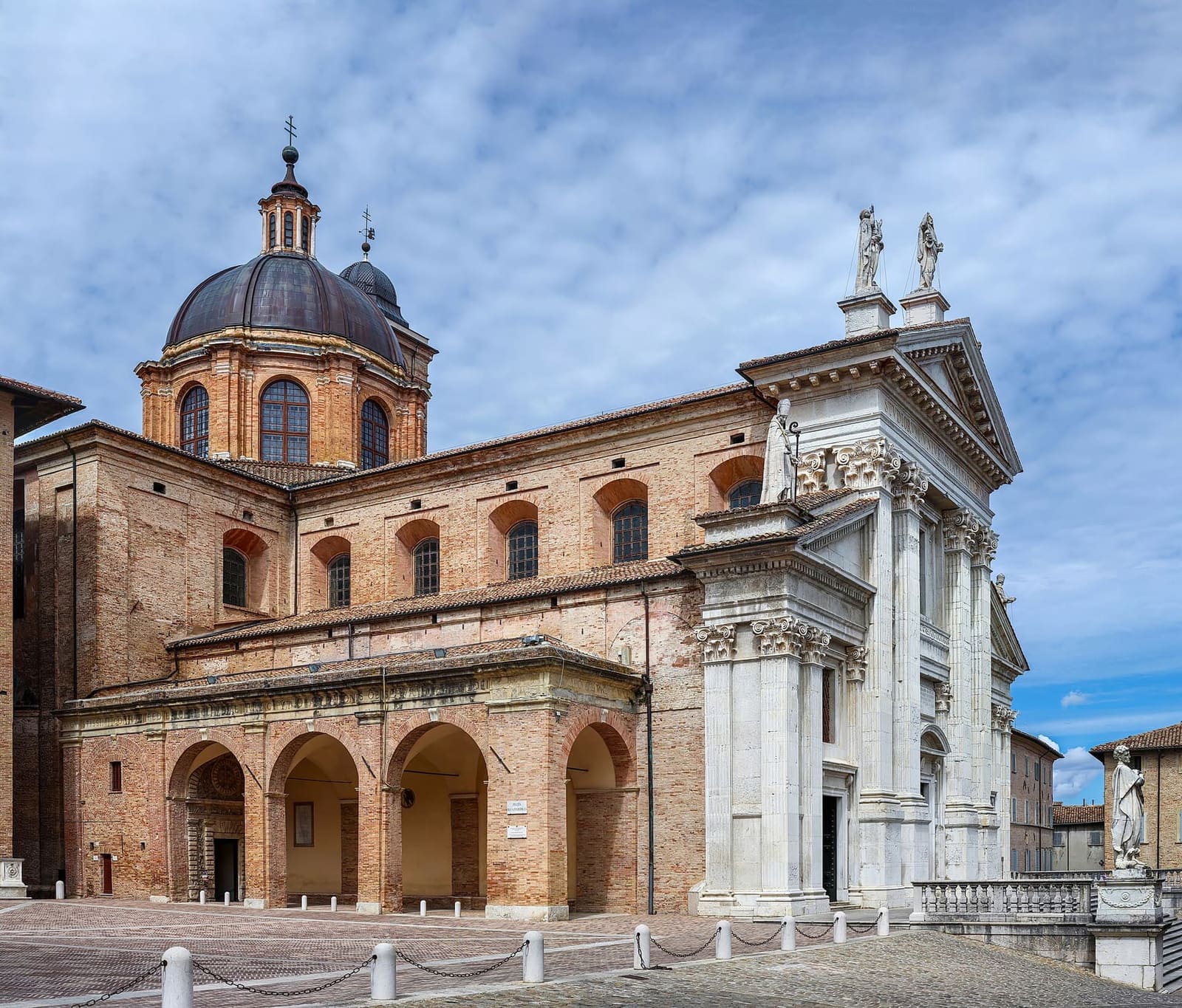 il duomo sulla piazza, con la facciata neoclassica di pietra bianca, il fianco di mattoni con il portico e la cupola sopra