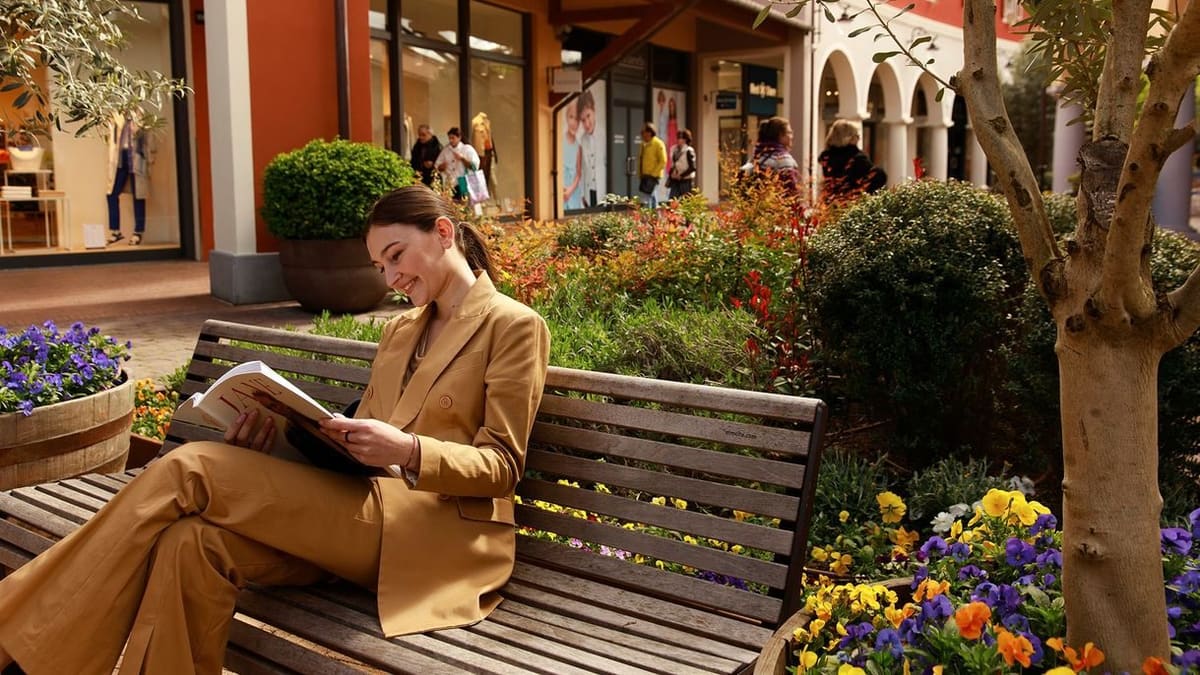 una visitatrice seduta a leggere sulla panchina del viale dei negozi, fra aiuole fiorite e olivi