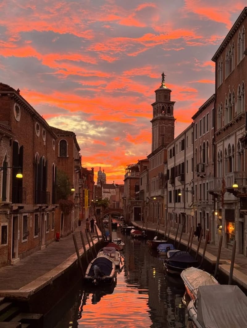 un rio stretto con le barche ormeggiate ai pali e il cielo rosso acceso sopra un campanile